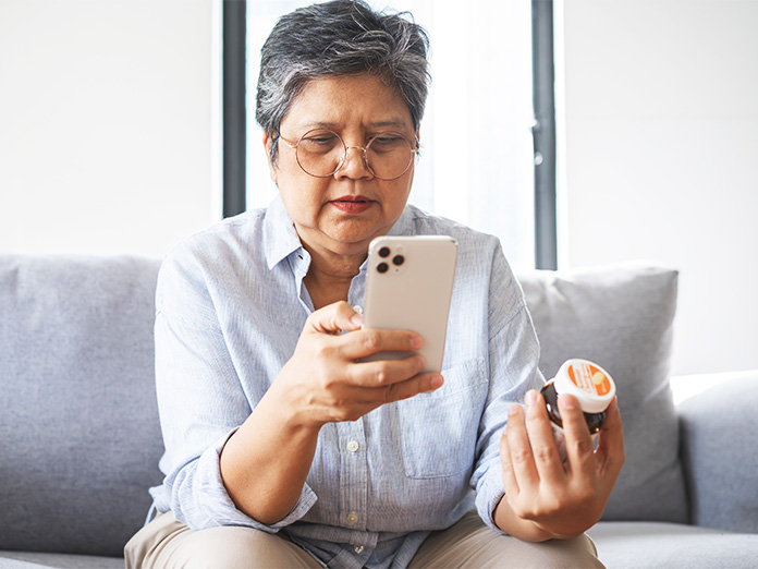An adult sitting on a couch at home uses a smartphone while holding a prescription medication bottle.